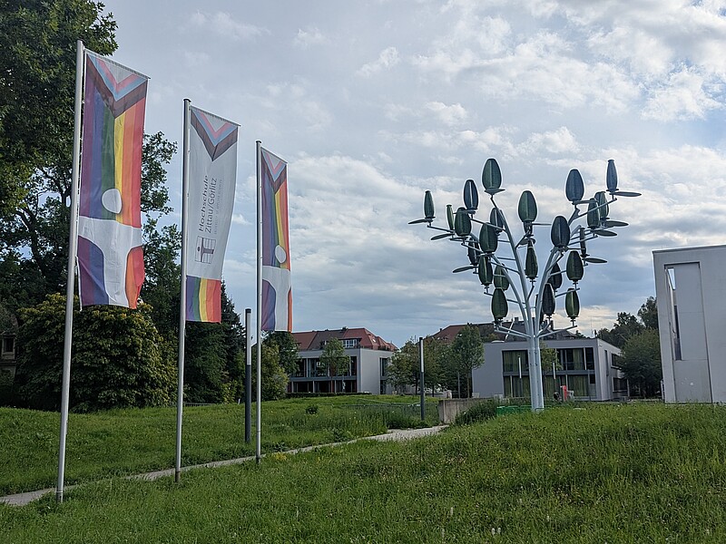 Vor dem Peter-Dierich-Haus am Zittauer Campus der Hochschule Zittau/Görlitz wehen Regenbogenfahnen neben dem erst vor kurzen installierten Wind-PV-Baum.